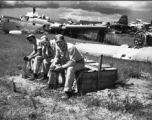 Three American GIs sitting on a discarded latrine box in front of wrecked American warplanes (including a B-24) in a boneyard at the base. An aircraft tail #349752 is in the background. Note men walking on wing of aircraft in background, undoubtedly engaged in salvage on the wing. In Yunnan, China, during WWII.