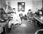 American photographers at work in the workshop in China during WWII. On the left Eugene T. Wozniak repairs a piece of photographic equipment.