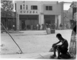 A tired looking young man sits in the opening to a stall advertising car parts, in Kunming, China, during WWII.