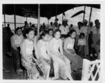 Local people in Burma near the 797th Engineer Forestry Company--Ladies in their finery sit under a covering, with a Union Jack flying in the background, in Burma.  During WWII.