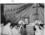 A parachute forms a backdrop in this Chapel in Kunming in 1945. Father Charles F. Meeus celebrates mass.  The Father's pulpit is the cowling for the P-40K "Little Flower" flown by Ray Kaiser (25th FS). "Little Flower" was St Therese from Lisieux, France. (Thanks JBarbaud)  Notice the corrugated metal making up the lower area of the alter, also likely parts from aircraft or packaging munitions.