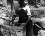 A young woman uses a wooden rod to husk rice in a large stone mortar. Local people in China, probably in Yunnan province.   From the collection of Wozniak, combat photographer for the 491st Bomb Squadron, in the CBI.