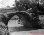 A rural woman shouldering baskets crosses a worn footbridge in China.  From the collection of Eugene T. Wozniak.