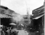 A very narrow main market street in a typical village near Kunming, Yunnan province. In addition to food stalls and other sellers, in this case a funeral activity (usually several days long) is being carried out in part in the street. During WWII.